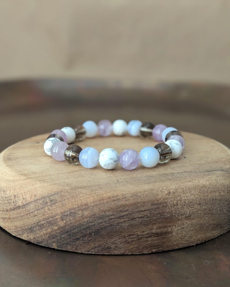 May include: A beaded bracelet featuring a repeating pattern of round, polished stones in shades of white with grey veining, translucent light blue, pale lavender, and dark smoky brown, all strung together on a flexible, circular elastic cord.