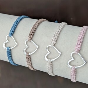 May include: Four macrame bracelets with silver heart charms. The bracelets are in shades of blue, tan, cream, and pink. The heart charms are centered on the bracelets.