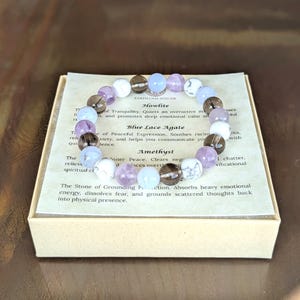 May include: A beaded bracelet featuring alternating round stones in shades of white, translucent light blue, smoky brown, and pale purple. The bracelet rests on a square card printed with the text Earth Gem Atelier, Howlite, Blue Lace Agate, and Amethyst.