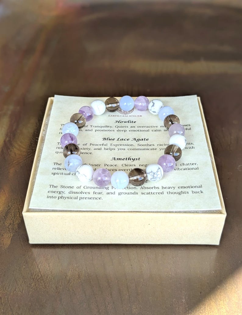 May include: A beaded bracelet featuring alternating round stones in shades of white, translucent light blue, smoky brown, and pale purple. The bracelet rests on a square card printed with the text Earth Gem Atelier, Howlite, Blue Lace Agate, and Amethyst.