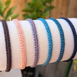 May include: A collection of six macrame bracelets in various colors, including brown, peach, lavender, and shades of blue. The bracelets are displayed on a white cylindrical form, showcasing their woven texture and design. The background features greenery.