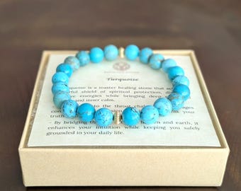 May include: A circular beaded bracelet made of bright blue turquoise stones with dark veining, featuring two small metallic spacer beads. The bracelet rests on a square card inside a box, with text describing the stone's properties visible.