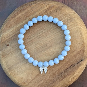 Puede incluir: Una pulsera de cuentas azul claro con un dije plateado de alas de &aacute;ngel. La pulsera est&aacute; hecha de cuentas redondas y lisas y se exhibe sobre una superficie de madera. Es una pieza de joyer&iacute;a.