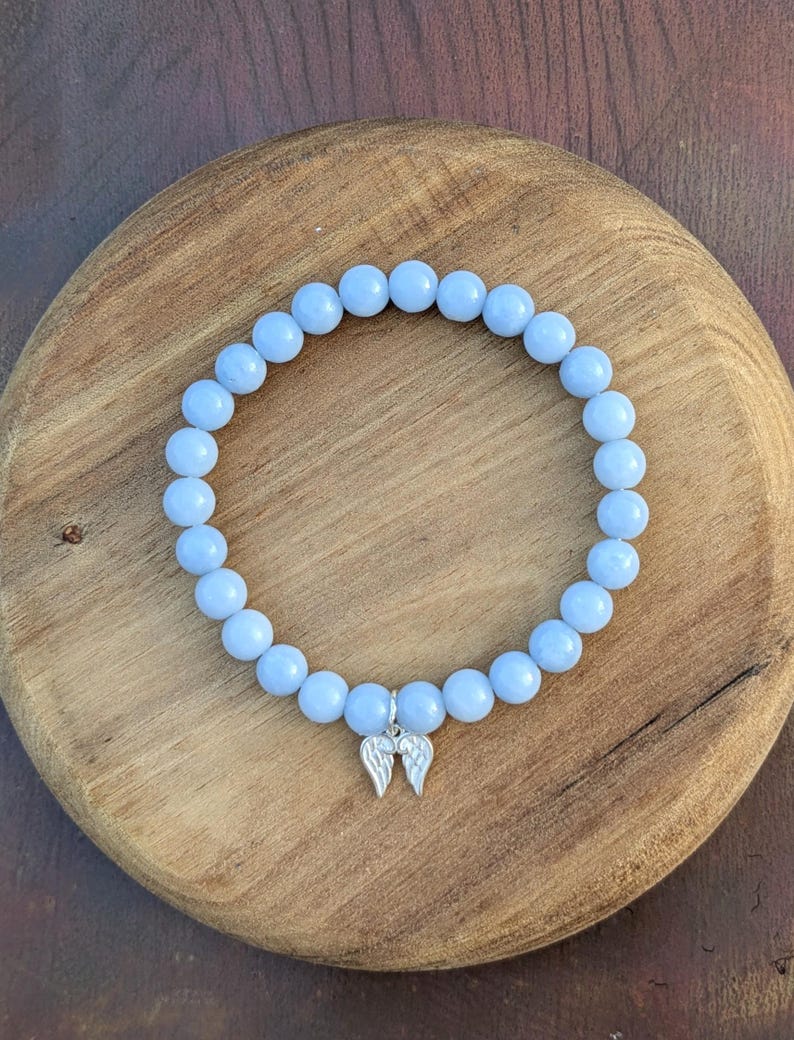 Puede incluir: Una pulsera de cuentas azul claro con un dije plateado de alas de &aacute;ngel. La pulsera est&aacute; hecha de cuentas redondas y lisas y se exhibe sobre una superficie de madera. Es una pieza de joyer&iacute;a.