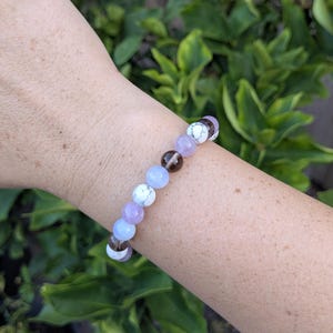 May include: A beaded bracelet featuring a repeating pattern of round, polished stones in shades of white with grey veining, translucent light blue, pale lavender, and dark brown, strung together on a flexible cord to form a circular band.