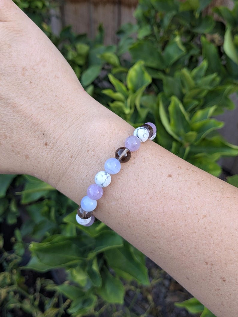 May include: A beaded bracelet featuring a repeating pattern of round, polished stones in shades of white with grey veining, translucent light blue, pale lavender, and dark brown, strung together on a flexible cord to form a circular band.