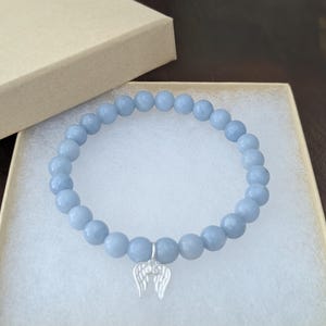 Puede incluir: Pulsera de cuentas azul claro con un dije de alas plateadas, presentada en una caja de regalo beige. La pulsera presenta cuentas redondas y lisas. La caja est&aacute; abierta, revelando la pulsera en su interior.