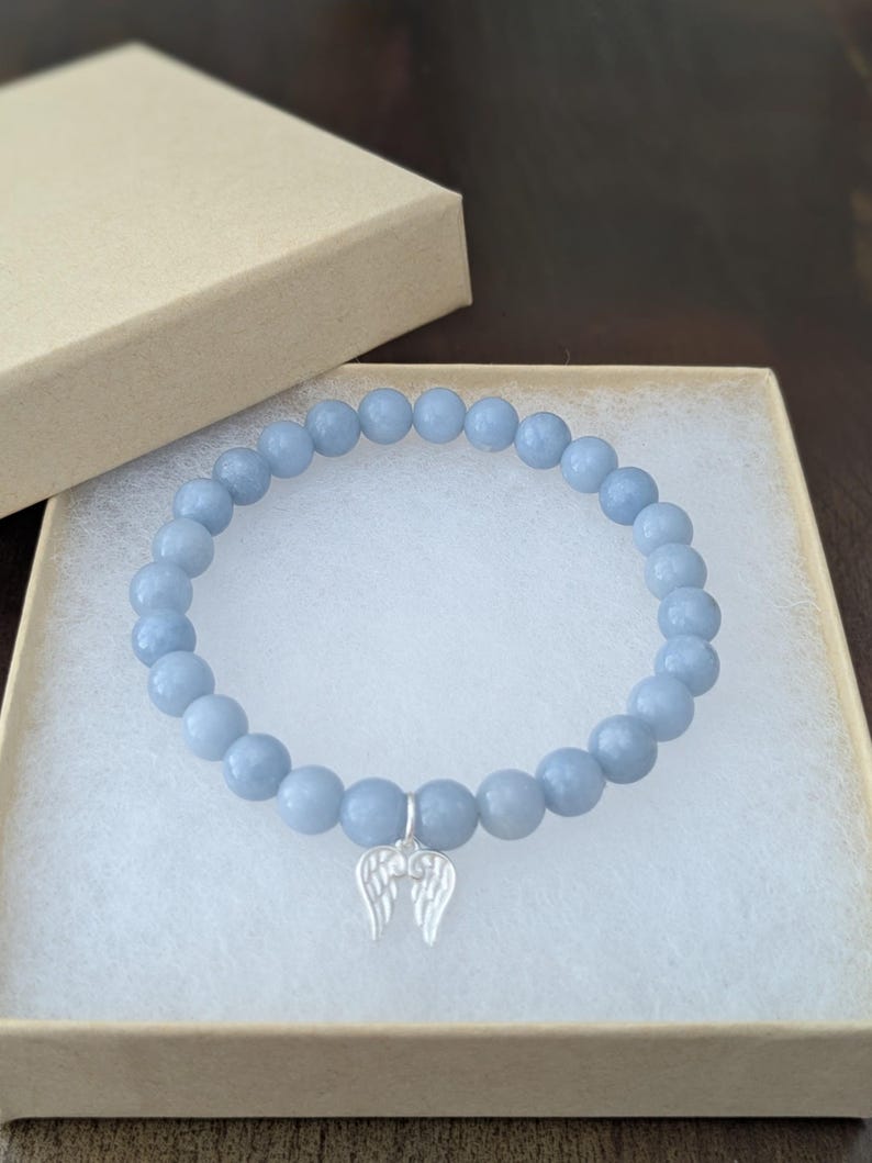Puede incluir: Pulsera de cuentas azul claro con un dije de alas plateadas, presentada en una caja de regalo beige. La pulsera presenta cuentas redondas y lisas. La caja est&aacute; abierta, revelando la pulsera en su interior.