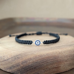 May include: A black cord bracelet featuring a central circular charm. The charm is designed with a white center surrounded by a ring of small blue stones, creating an eye-like pattern. The bracelet has a braided texture and adjustable cord ends.