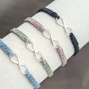 May include: Four macrame bracelets in blue, green, pink, and black. Each bracelet has a silver infinity symbol. The bracelets are displayed on a white surface, showcasing the different colours and designs.