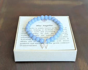 May include: A light blue beaded bracelet made of smooth, round stones. The bracelet features a small, silver-toned metal charm shaped like a pair of angel wings. It is displayed on a square white card with printed text about Blue Angelite.