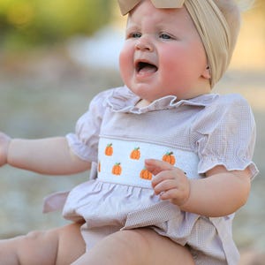 May include: A beige gingham romper featuring a white smocked chest panel with embroidered orange pumpkins, short puff sleeves with ruffled collars, and a matching beige fabric hair bow headband worn on the head.