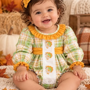 May include: A long-sleeved romper featuring a green, white, and orange plaid pattern with a mustard yellow ruffled collar and sleeve cuffs. A white vertical center panel displays three embroidered turkey motifs and a single metal snap closure.
