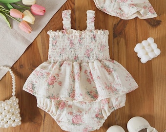 May include: A cream-colored romper with a pink and green floral pattern, featuring a smocked bodice, ruffled waist, and shoulder straps. The set includes a matching bucket hat with a floral print and a pair of white shoes with lace trim.