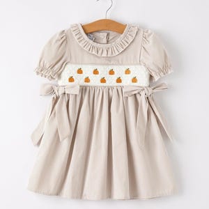 May include: A beige gingham dress featuring short puff sleeves, a ruffled collar, and a white smocked bodice embroidered with small orange pumpkins. The dress includes side waist ties that form bows and a gathered skirt, hanging on a wooden hanger.