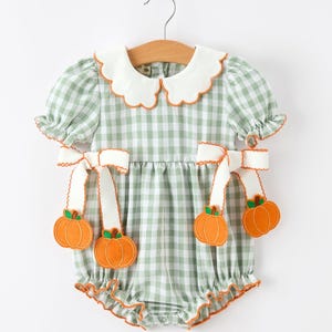 May include: A sage green and white gingham romper featuring a white scalloped collar with orange trim, short puffed sleeves, and white ribbon ties at the waist. Four orange pumpkin appliqués hang from the ribbons, and the leg openings are ruffled.