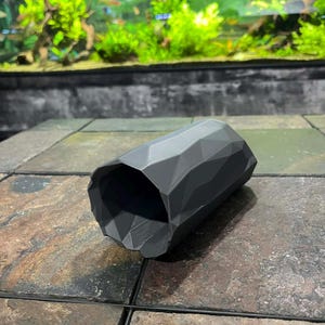May include: A dark grey, geometric aquarium decoration. The cylindrical structure has a faceted design, resembling a rock formation. The background shows an aquarium with plants and fish.
