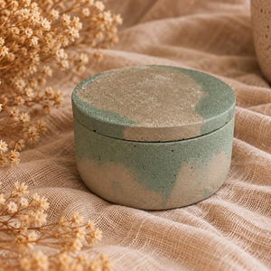May include: A round, lidded container made of concrete with a marbled finish in shades of sage green and beige. The cylindrical vessel features a flat, circular lid that sits flush with the base, showcasing a textured, porous surface.