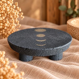 May include: A circular, dark gray, textured pedestal tray with three short legs. The top surface features a gold-colored graphic depicting the phases of the moon, arranged in a vertical line from a full circle to a thin crescent.