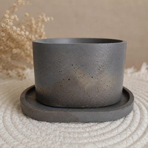 May include: A dark gray cylindrical concrete planter with a porous, textured surface and a subtle metallic bronze gradient finish near the base, resting on a matching circular concrete saucer tray with a smooth, flat rim and a solid base.