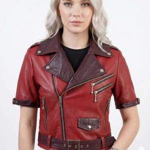 May include: A red leather jacket with short sleeves and a burgundy collar, lapels, and accents. The jacket features a zipper closure, multiple pockets, and a belt at the waist. Silver hardware is visible.