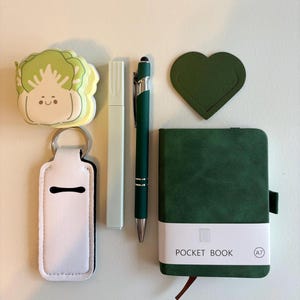 May include: An assortment of stationery items including a green pen, a white highlighter, a small green notebook with the text "POCKET BOOK", a green heart-shaped bookmark, a white keychain, and a cabbage-shaped sticker.