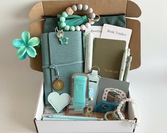 May include: A cardboard gift box containing a teal notebook with a charm, a green pouch, a beaded keychain, a small olive green To-Do List notebook, a Reading Book Tracker card, pens, a heart-shaped tag, a teal clip, and various stationery items.
