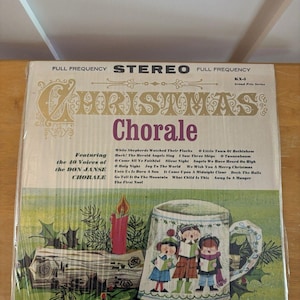 Puede incluir: Una portada de disco de vinilo titulada Christmas Chorale con el Don Janse Chorale. La portada muestra un tronco de Navidad con una vela roja y una taza ilustrada con tres cantantes. La lista de canciones incluye While Shepherds Watched.