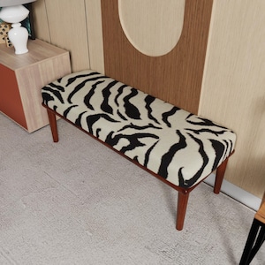 May include: A rectangular bench featuring a black and white zebra print fabric upholstery on the seat. The bench is supported by four tapered wooden legs in a medium brown finish, providing a stable base for the padded seating surface.