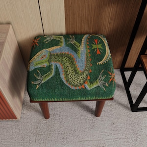 May include: A small, square footstool with a green fabric top featuring a detailed lizard design in shades of green, blue, and orange. The footstool has four short, dark wooden legs, and is suitable for a living room or bedroom.