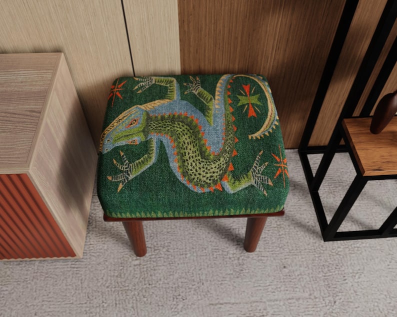 May include: A small, square footstool with a green fabric top featuring a detailed lizard design in shades of green, blue, and orange. The footstool has four short, dark wooden legs, and is suitable for a living room or bedroom.
