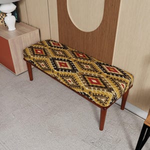 May include: A rectangular bench with a patterned seat cushion. The cushion features a repeating geometric design in shades of yellow, red, black, and brown. The bench has four wooden legs.