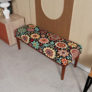 May include: A wooden bench with a floral patterned cushion. The cushion features a black background with colorful flowers in shades of red, yellow, blue, and orange. The bench has four brown legs.