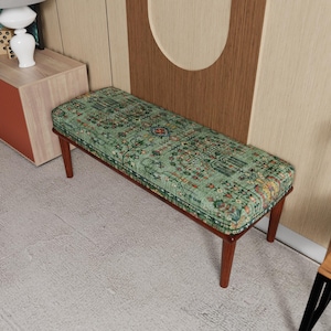 Op de afbeelding: Een rechthoekige bank met een groen kussen met patroon en donkerbruine houten poten. Het kussen heeft een gedetailleerd ontwerp met verschillende kleuren. De bank is een meubelstuk, geschikt voor een woonkamer of slaapkamer.