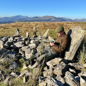 Puede incluir: Una persona con chaqueta de lana a cuadros marr&oacute;n, pantalones verde oscuro, botas de goma verde oscuro y gorra plana, sentada sobre una gran losa de piedra en una estructura circular, sosteniendo un cuaderno y un bol&iacute;grafo verde en las manos.