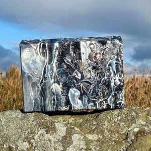 May include: A rectangular block featuring a detailed black and white illustration with numerous overlapping figures, including human faces, animals, and celestial elements, rendered in a high-contrast style with intricate white line work on a dark background.