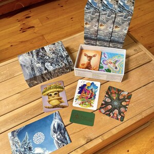 Puede incluir: Un juego de cartas en caja que incluye tres cajas verticales altas con im&aacute;genes azules y blancas, una tapa de caja rectangular con arte abstracto oscuro y una caja abierta que contiene cartas con ilustraciones de un ciervo, un colibr&iacute;, un b&uacute;ho y personas.