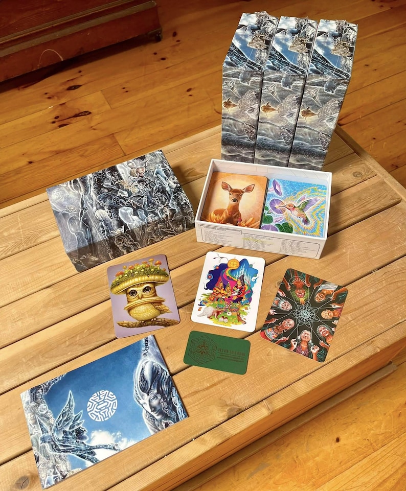 Puede incluir: Un juego de cartas en caja que incluye tres cajas verticales altas con im&aacute;genes azules y blancas, una tapa de caja rectangular con arte abstracto oscuro y una caja abierta que contiene cartas con ilustraciones de un ciervo, un colibr&iacute;, un b&uacute;ho y personas.