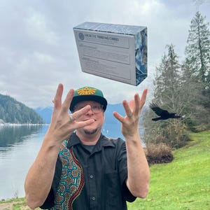 Puede incluir: Una caja rectangular etiquetada como Galactic Trading Cards con un patr&oacute;n de m&aacute;rmol azul y blanco, flotando en el aire sobre una persona que viste una camisa negra de manga corta con un patr&oacute;n geom&eacute;trico colorido y una gorra de b&eacute;isbol verde.