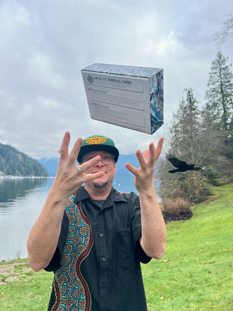 Puede incluir: Una caja rectangular etiquetada como Galactic Trading Cards con un patr&oacute;n de m&aacute;rmol azul y blanco, flotando en el aire sobre una persona que viste una camisa negra de manga corta con un patr&oacute;n geom&eacute;trico colorido y una gorra de b&eacute;isbol verde.