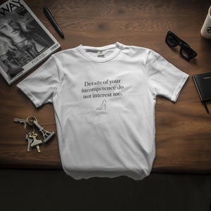 Può includere: T-shirt bianca con la scritta "Details of your incompetence do not interest me." e un disegno di scarpa con tacco alto. La maglietta è appoggiata su una superficie di legno scuro, insieme a una rivista, occhiali da sole, un piccolo taccuino, chiavi, una penna e una tazza di caffè.