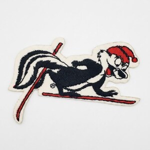 May include: An embroidered patch featuring a black and white skunk wearing a red Santa hat, depicted in a skiing pose with red skis on a white background. The skunk has a bushy tail and is shown in profile, with red lines representing ski poles.