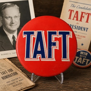 May include: A circular red political campaign button featuring the word TAFT in large blue letters with a white outline. The button is displayed on a clear plastic stand, positioned in front of historical campaign literature and a portrait of Robert A. Taft.