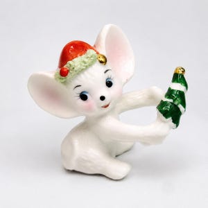 Puede incluir: Una figura de ratón de cerámica blanca con orejas grandes, mejillas rosadas y ojos azules. El ratón lleva un gorro de Papá Noel rojo con un borde verde texturizado y sostiene un pequeño adorno de árbol de Navidad verde y blanco.