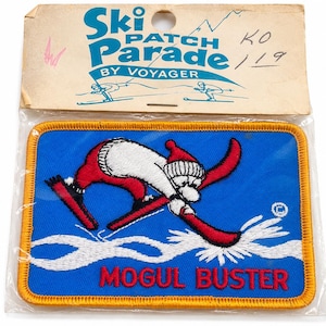 Op de afbeelding: Een rechthoekige geborduurde patch met een blauwe achtergrond en een gele rand. Het ontwerp toont een witte beer met een rode muts en rode broek, skiënd op witte sneeuw. De tekst MOGUL BUSTER is in het rood. De koptekst luidt Ski Patch Parade.