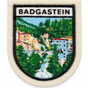 May include: A shield-shaped embroidered patch with a cream-coloured border. The centre features a colourful landscape scene of buildings, trees, and a waterfall, with the text BADGASTEIN printed in black capital letters on a white background at the top.