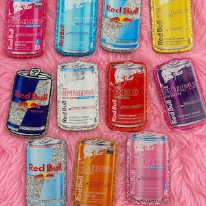May include: A collection of eleven rectangular patches shaped like Red Bull energy drink cans in various colors including pink, blue, yellow, red, and orange. Each patch features a glittery border and printed text detailing specific flavor editions.