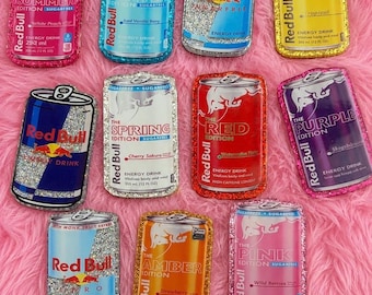 Puede incluir: Una colección de once parches rectangulares con forma de latas de bebida energética Red Bull en varios colores, incluyendo rosa, azul, amarillo, rojo y naranja. Cada parche presenta un borde brillante y texto impreso con las ediciones.