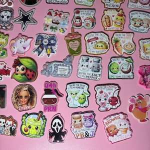 Puede incluir: Una colección de pines de acrílico con purpurina que presentan varios personajes de dibujos animados, parejas de alimentos con juegos de palabras, logotipos deportivos e iconos de la cultura pop. Cada pin tiene una forma y diseño único.