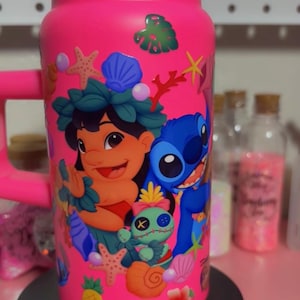 May include: A bright pink tumbler featuring a colorful design of Lilo and Stitch characters, along with tropical elements like seashells, starfish, and flowers. The tumbler has a red rim and handle, and is decorated with various stickers.